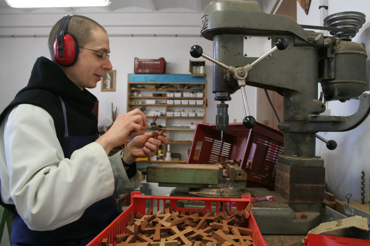 Un frère, casque antibruit sur les oreilles, perce de petites croix de bois devant une caisse qui en est remplie